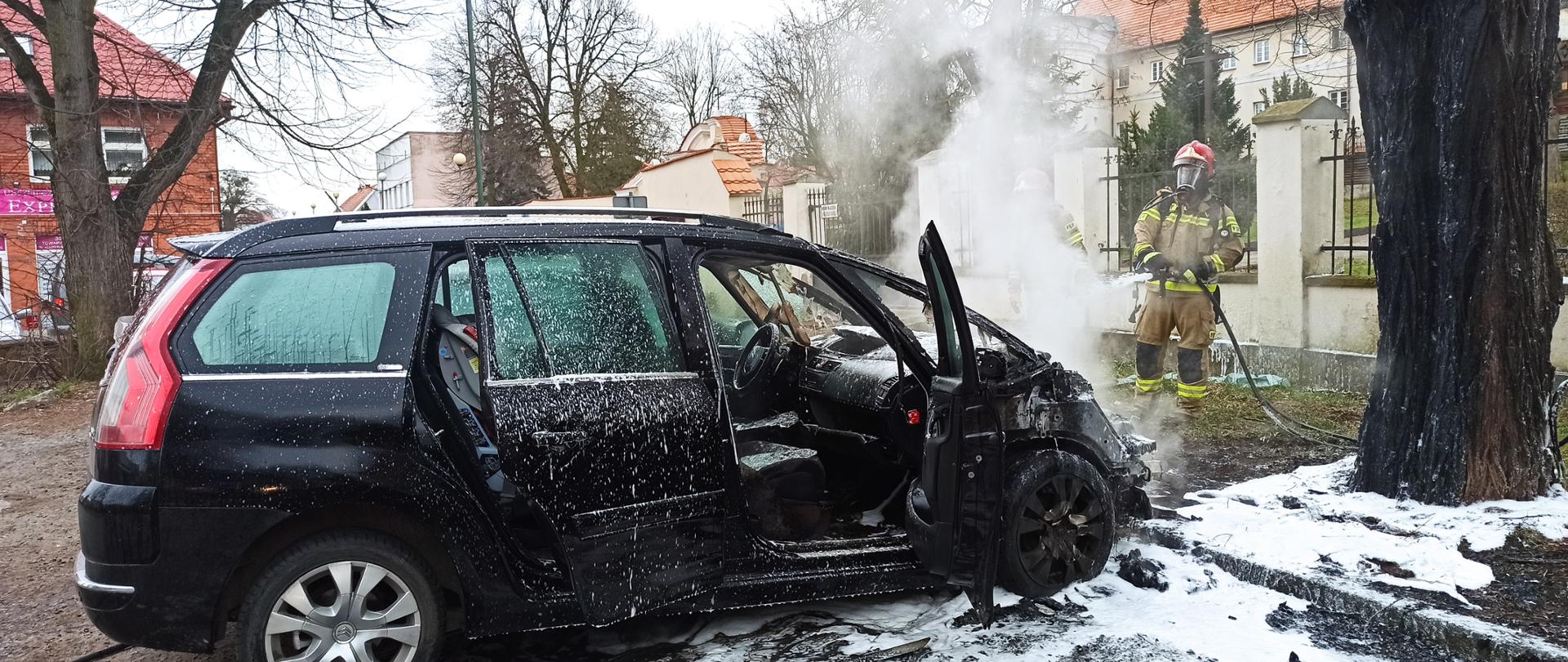 Zdjęcie przedstawia zaparkowany samochód osobowy na parkingu, który uległ zapaleniu. Pożarem objęta komora silnika i deska rozdzielcza. Strażak podaje środek gaśniczy na palący się pojazd. 