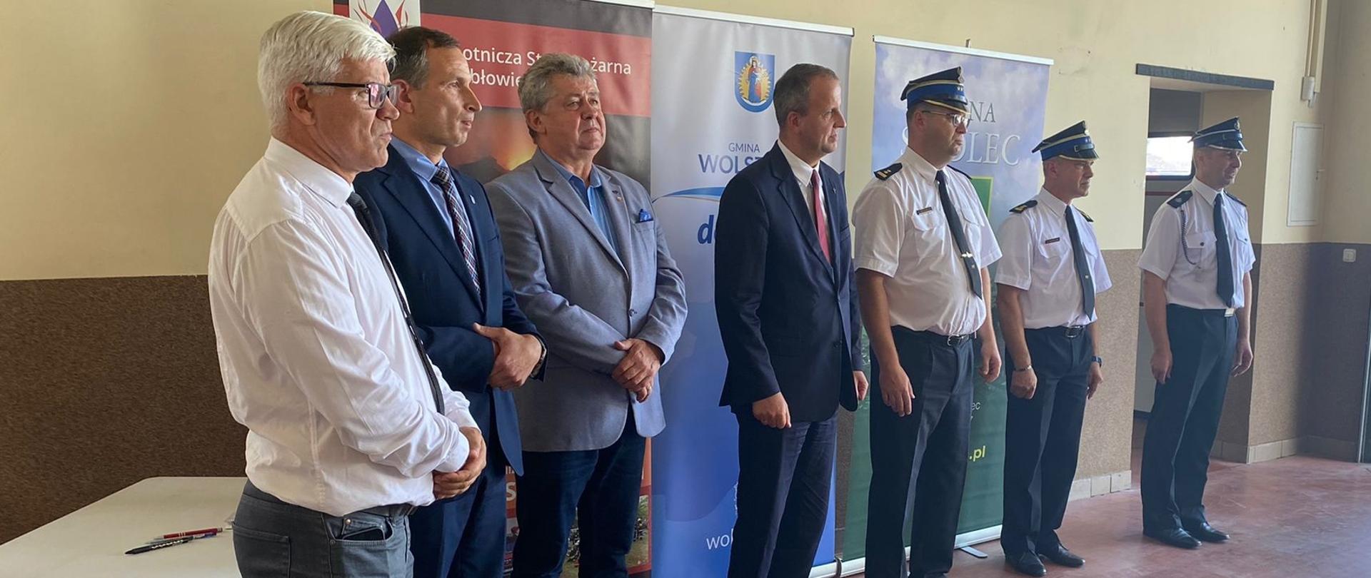 Zdjęcie wykonane w siedzibie OSP Kębłowo. Na pierwszym planie stoją zaproszeni przedstawiciele władz: od lewej: Pan Jacek Kolesiński – Wójt Gminy Siedlec, Pan Tomasz Spiralski - Z-ca Burmistrza Wolsztyna, Pan Jacek Skrobisz – Starosta Wolsztyński, Pan Michał Zieliński - Wojewoda Wielkopolski, bryg. Robert Natunewicz - Zastępca Wielkopolskiego Komendanta Wojewódzkiego PSP, bryg. Roman Rembielak - Komendant Powiatowy Państwowej Straży Pożarnej w Wolsztynie, druh Michał Nowak - przedstawiciel OSP Kębłowo. W tle widoczne banery z napisem Gmina Siedlec, Gmina Wolsztyn oraz Ochotnicza Straż Pożarna Kębłowo.