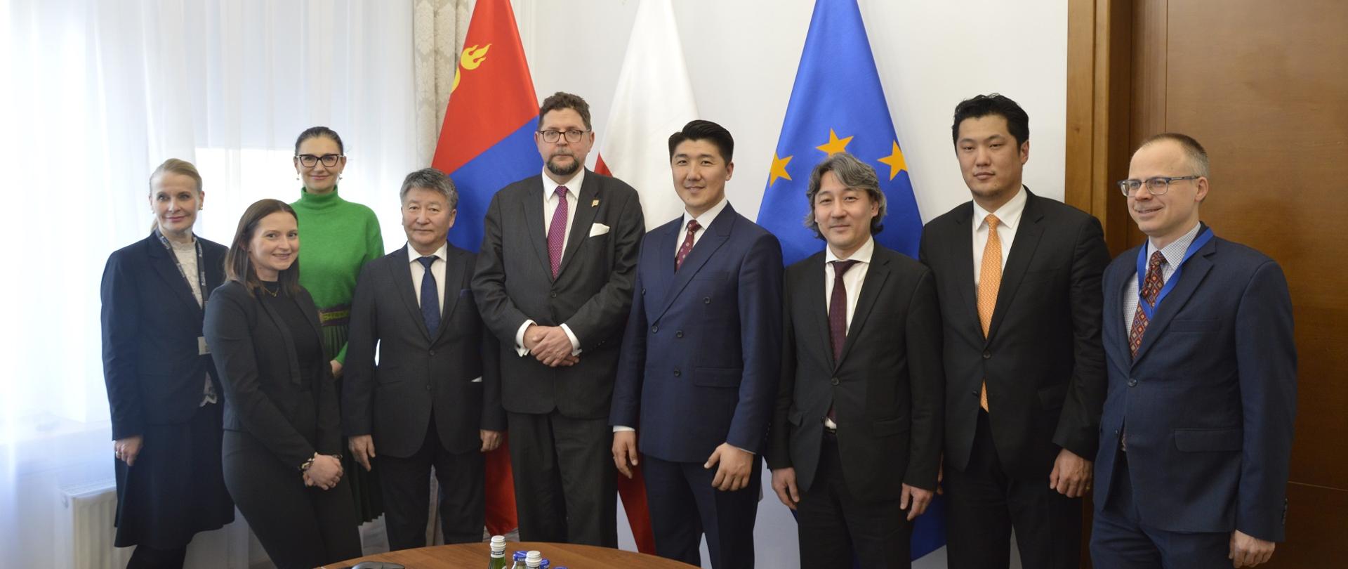 Pod ścianą sali stoi grupa dziewięciu osób, pośrodku wiceminister Szeptycki, za nimi flagi Polski, Mongolii i UE.