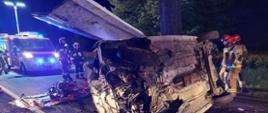 Na zdjęciu widać zniszczony samochód osobowy lecący na boku, na drugim planie widać strażaków, na ulicy leży sprzęt hydrauliczny oraz w tle 