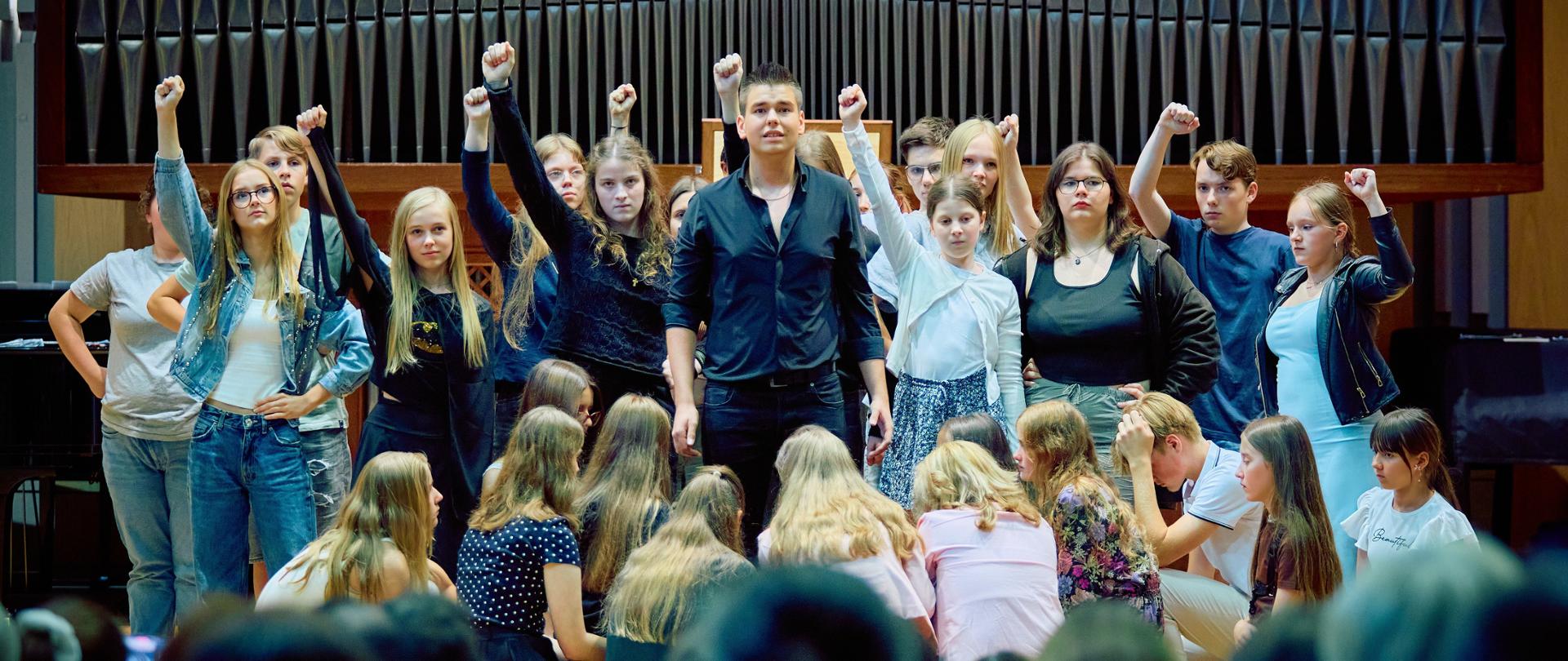 Zdjęcie przedstawia grupę nastolatków stojących na scenie z zaciśniętymi pięściami uniesionymi w górę, co sugeruje gest siły, jedności lub protestu. Poniżej nich, bliżej pierwszego planu, siedzi inna grupa nastolatków, zwróconych plecami do widza. Całość dzieje się w dużej sali koncertowej z dużymi piszczałkami organowymi w tle, co dodaje miejscu podniosłego charakteru. Na samym dole, rozmyte, widać z tyłu głowy publiczności. Oświetlenie sceny jest jasne, podczas gdy publiczność znajduje się w cieniu, co skupia uwagę na występujących.
