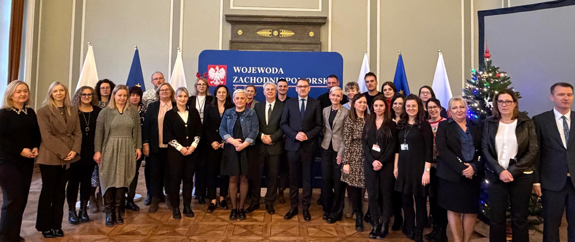 Grupowe zdjęcie kilkudziesięciu osób stojących w reprezentacyjnej sali urzędowej. W tle godło Polski na ścianie, flagi oraz baner „Wojewoda Zachodniopomorski”. Po prawej stronie widoczna udekorowana choinka. Uczestnicy ubrani formalnie, ustawieni w kilku rzędach, pozują do wspólnej fotografii.