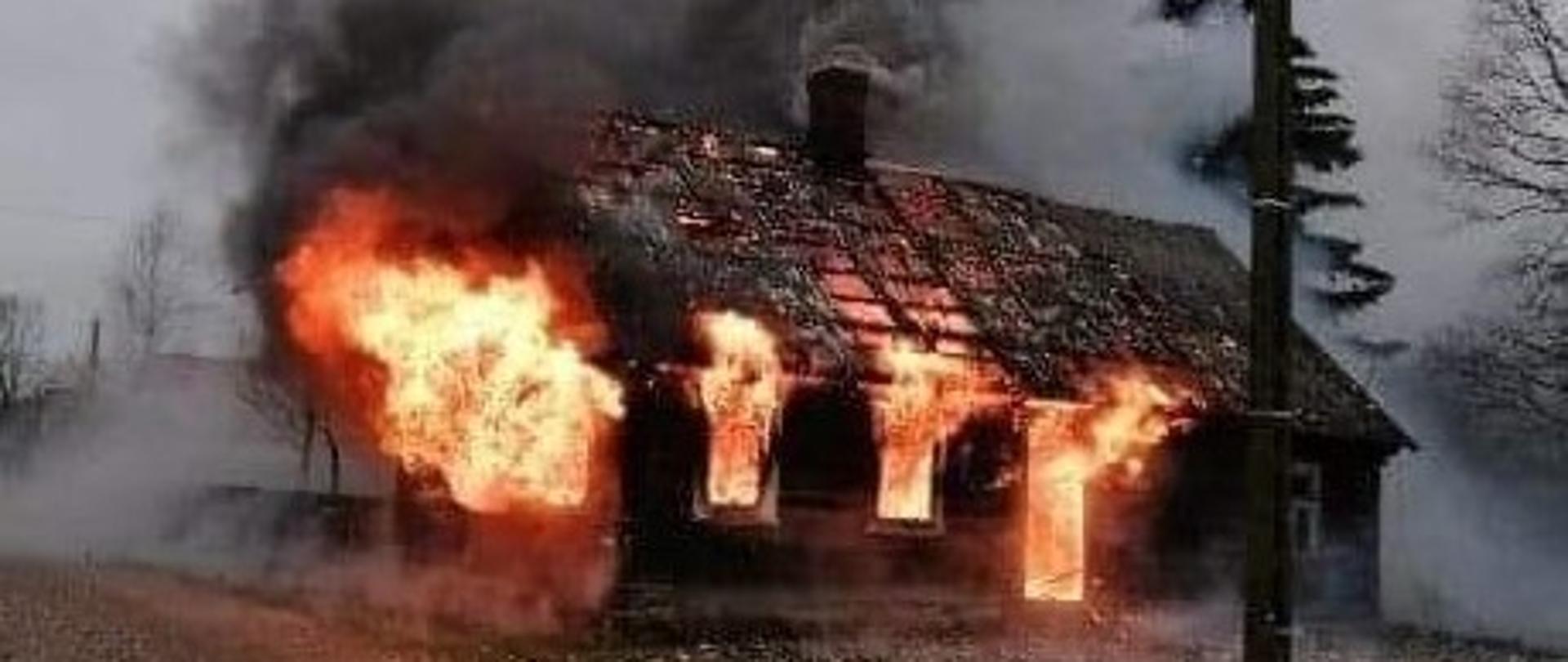 Palący się dom jednorodzinny. Pożarem objęty jest cały jednokondygnacyjny, drewniany budynek mieszkalny. Przez okna i drzwi wydostają się na zewnątrz płomienie. Część pokrycia dachu (eternit) jest zniszczona. Na poddaszu również widoczne są płomienie. Nad budynkiem unosi się gęsty dym.