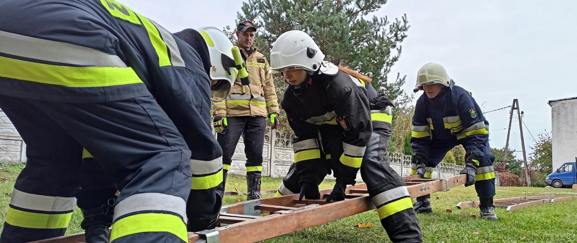 Strażacy sprawiają (składają) drabinę nasadkową. Zdjęcie wykonane z perspektywy podłoża. Pierwszy od lewej strażak ustawiony tyłem pochylony nad drabiną przytrzymuje pierwsze przęsło drabiny. Kolejna dwójka, pochylona nad kolejnym przęsłem ustawiona jeden za drugim wsuwa bocznice kolejnego przęsła w poprzednie. Za nimi ułożone na murawie, w ciągu jeszcze dwa przęsła drabiny. W tle nadzorujący ćwiczenie strażak PSP. 