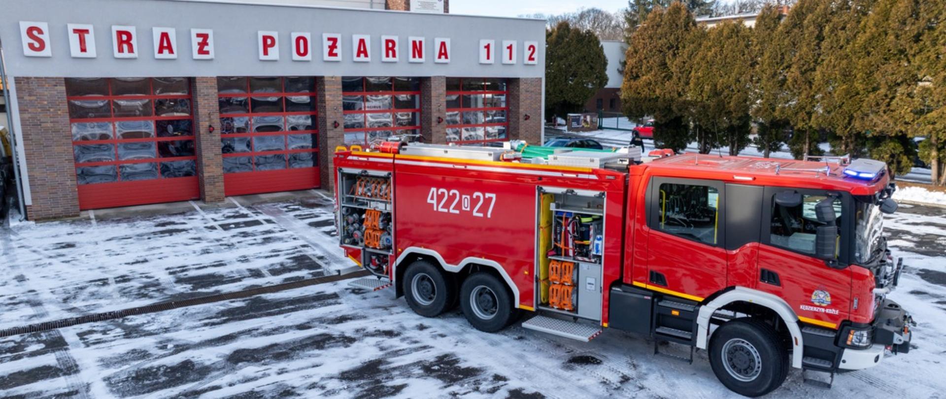 Samochód Scania P450 6x6 z otwartymi skrytkami sprzętowymi, prezentujący wyposażenie ratownicze, w tym węże, narzędzia i deski ratownicze. W tle budynek straży pożarnej „STRAŻ POŻARNA 112”.