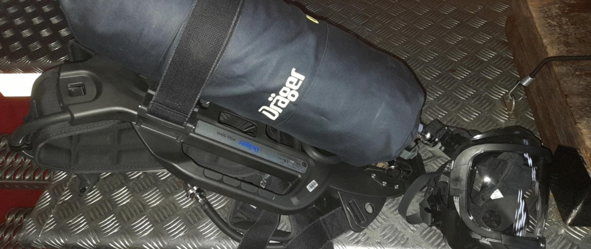 Na zdjęciu widoczny aparat powietrzny leżący na samochodzie pożarniczym - czarny noszak, butla w granatowym pokrowcu z napisem Drager oraz maska.