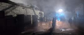 Na zdjęciu w porze nocnej widać na pierwszym planie strażaków w panującym zadymieniu przed budynkiem gospodarczym, w którym doszło do pożaru. W oddali samochód strażacki oświetlający teren akcji.
