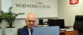 Wojewoda Lubuski w czasie seminarium on-line. Wojewoda siedzi przy stole w swoim gabinecie, na pierwszym planie otwarty laptop. W tle logo Wojewoda Lubuski