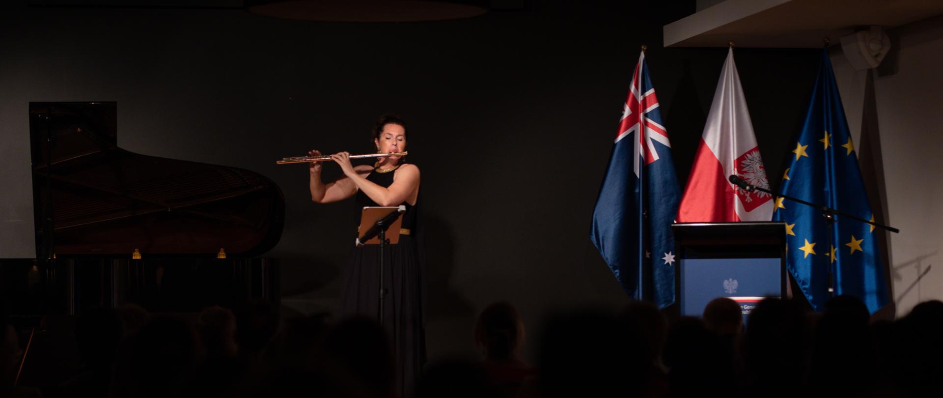 Flecistka Ewa Kowalski podczas występu na scenie, obok flagi państwowe. 