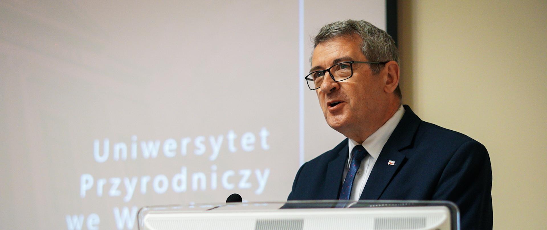 Minister przemawia stojąc za biurkiem, przed nim monitor komputerowy.