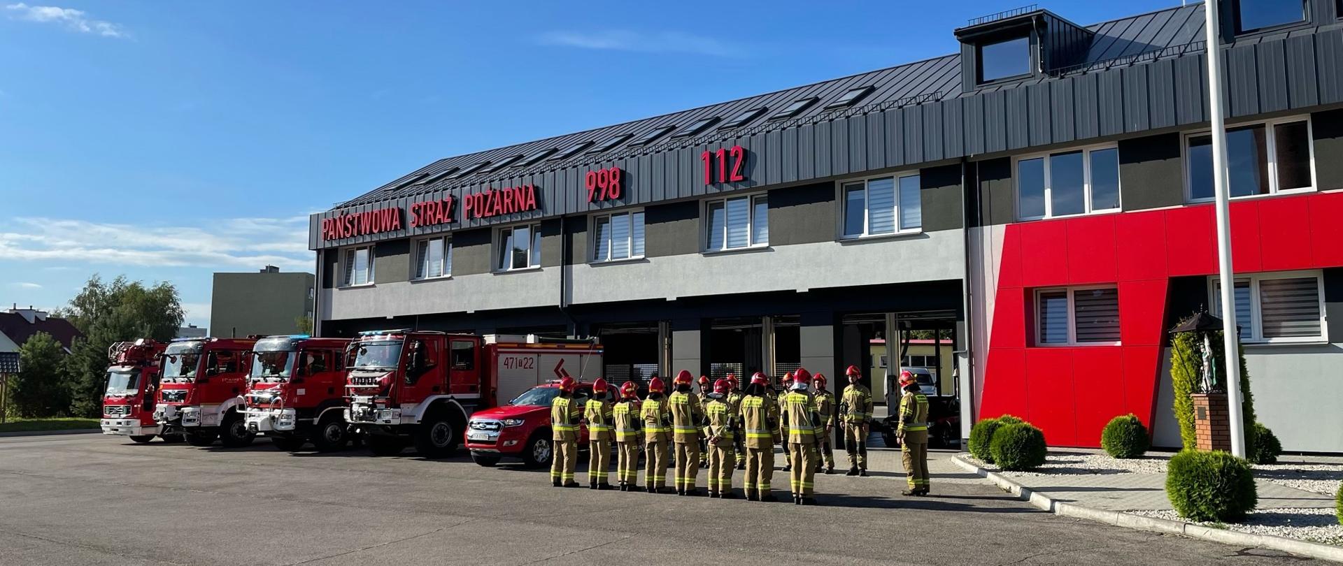 Na zdjęciu widać strażaków oddających hołd funkcjonariuszowi Komendy Głównej PSP, remizę straży pożarnej oraz samochody pożarnicze stojące po lewej stronie od strażaków przed remizą na włączonych sygnałach świetlno-dźwiękowych. Po prawej stronie stoi maszt z uniesioną flagą Polski.