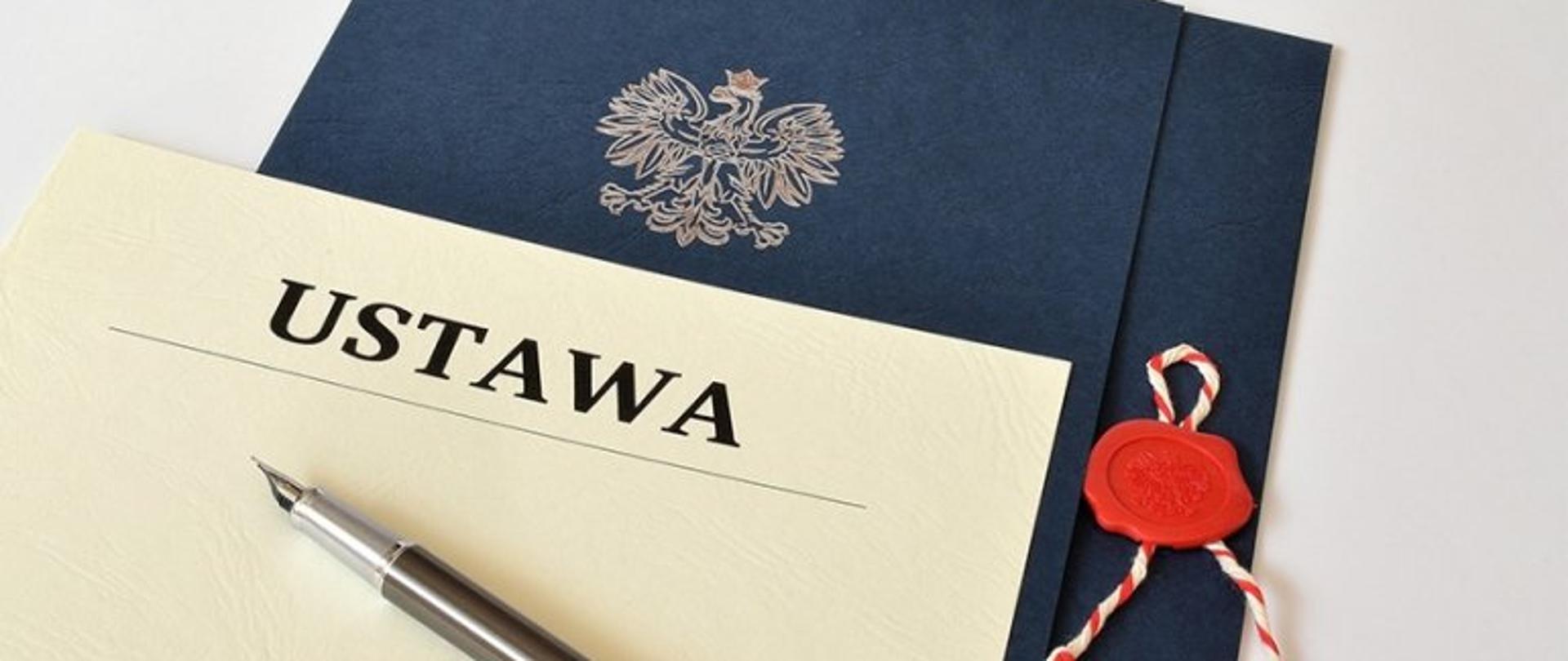 Niebieska teczka z godłem, na niej napis ustawa na kremowym tle oraz pióro wieczne.