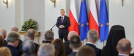 Widok z tyłu na salę, przed dużą grupą ludzi siedzących na krzesłach stoi prezydent Duda i mówi do mikrofonu na stojaku, za nim flagi Polski i UE, z boku na stoliku bukiet kwiatów.