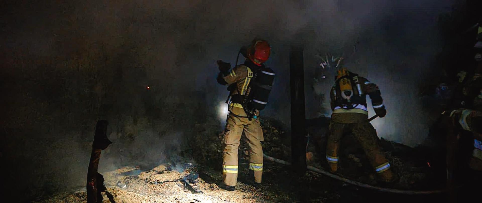 Strażacy prowadzą działania gaśnicze ubrani w umundurowanie strażackie i aparaty oddechowe. Obok nich panuje duże zadymienie. Zdjęcie wykonane w porze nocnej.