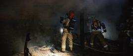 Strażacy prowadzą działania gaśnicze ubrani w umundurowanie strażackie i aparaty oddechowe. Obok nich panuje duże zadymienie. Zdjęcie wykonane w porze nocnej.