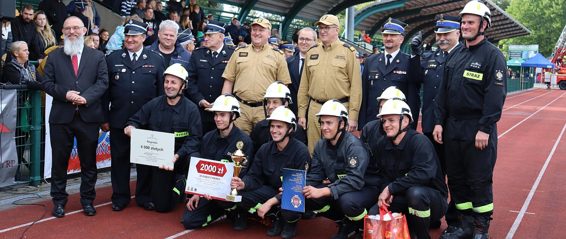 zwycięska drużyna ochotniczej straży pożarnej pozująca do zdjęcia na stadionie sportowym, wśród zawodników stoją również komendanci z państwowej straży pożarnej w tle zadaszenie trybun