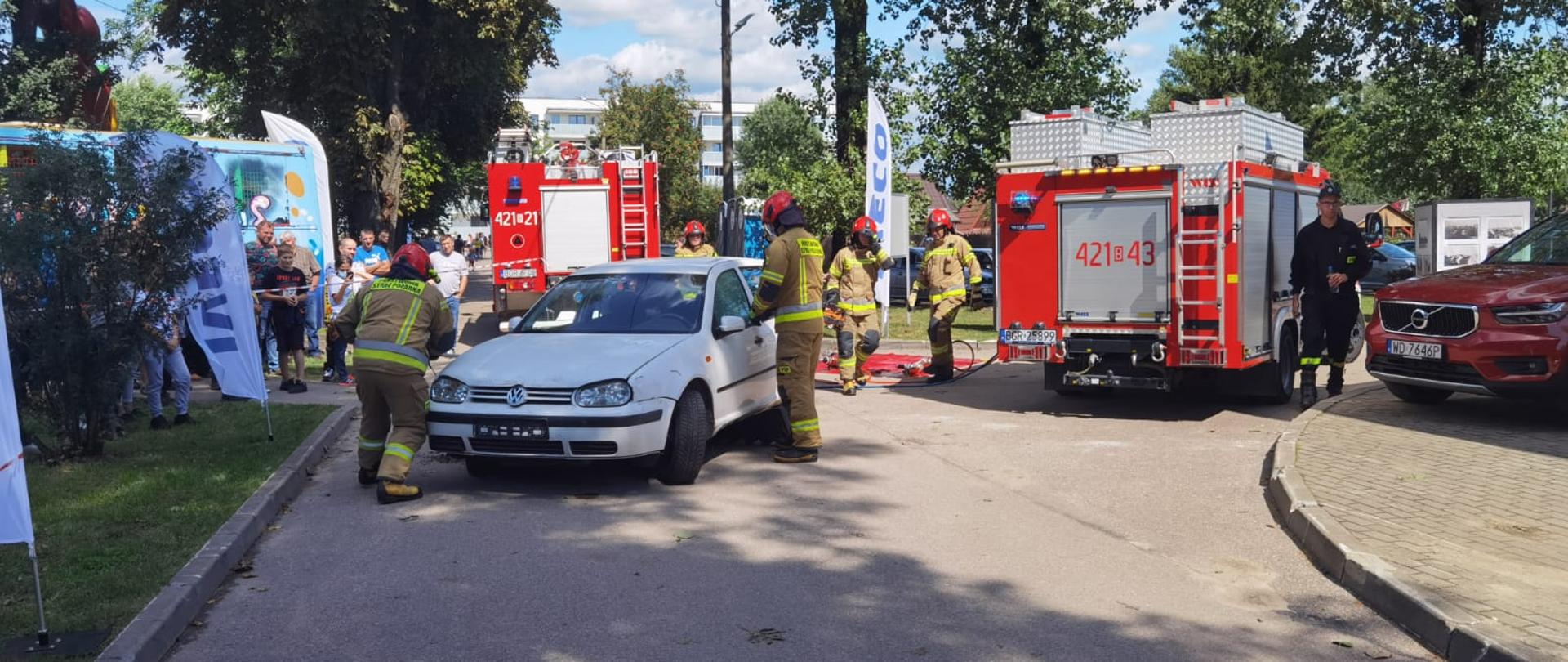 Samochód osobowy stojący na drodze przy nim strażacy PSP wykonują pokaz ratownictwa drogowego dla publiczności. W tle pojazdy straży pożarnej oraz publiczność.
