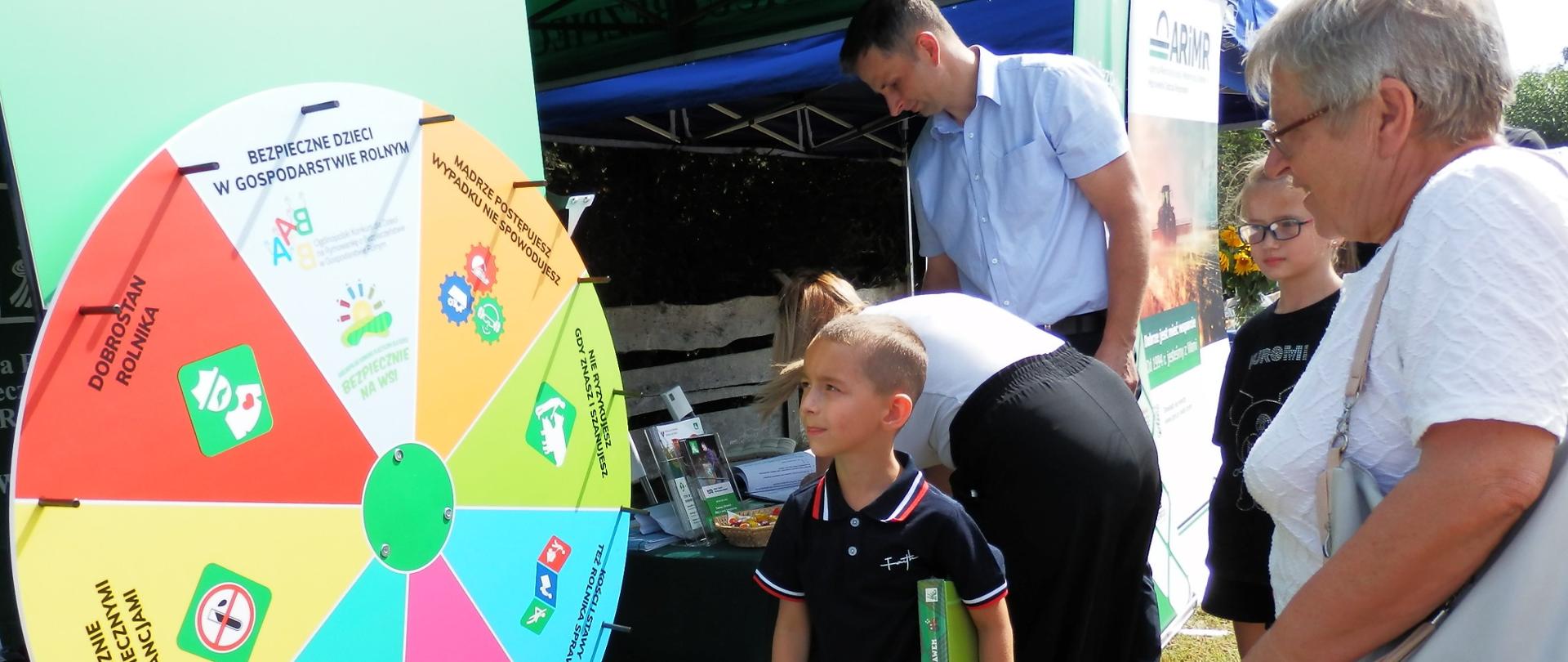 Przy namiocie KRUS stoi Koło Bezpieczeństwa Gdy Prewencyjnej, obok stoi chłopiec z babcią, który brał udział w grze i patrzy na koło. Za nimi pochylona przy stoliku kobieta rozwiązuje test w konkursie dla rolników.