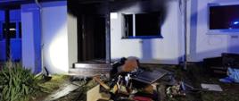 Noc. Widok na elewacje budynku mieszkaniowego, parterowego z płaskim dachem. Przednia elewacja w kolorze białym. Brak przeszkleń w oknie oraz drzwi. Okopcona elewacja nad otworem drzwiowym oraz oknem. Przed budynkiem, pod oknem leży sterta nadpalonych elementów wyposażenia mieszkania.
