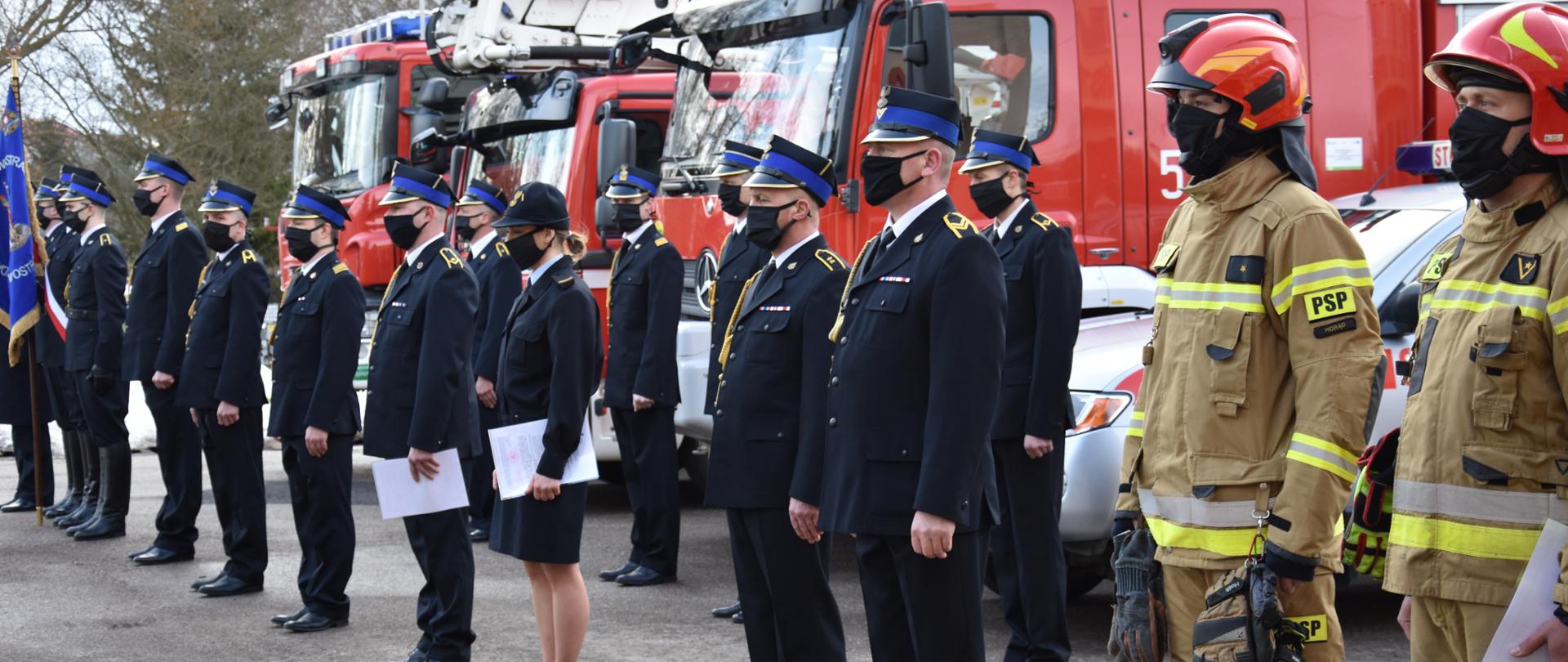W głównym kadrze funkcjonariusze odchodzący na zaopatrzenie emerytalne w mundurach wyjściowych. W tle widać pojazdy pożarnicze.