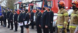 W głównym kadrze funkcjonariusze odchodzący na zaopatrzenie emerytalne w mundurach wyjściowych. W tle widać pojazdy pożarnicze.