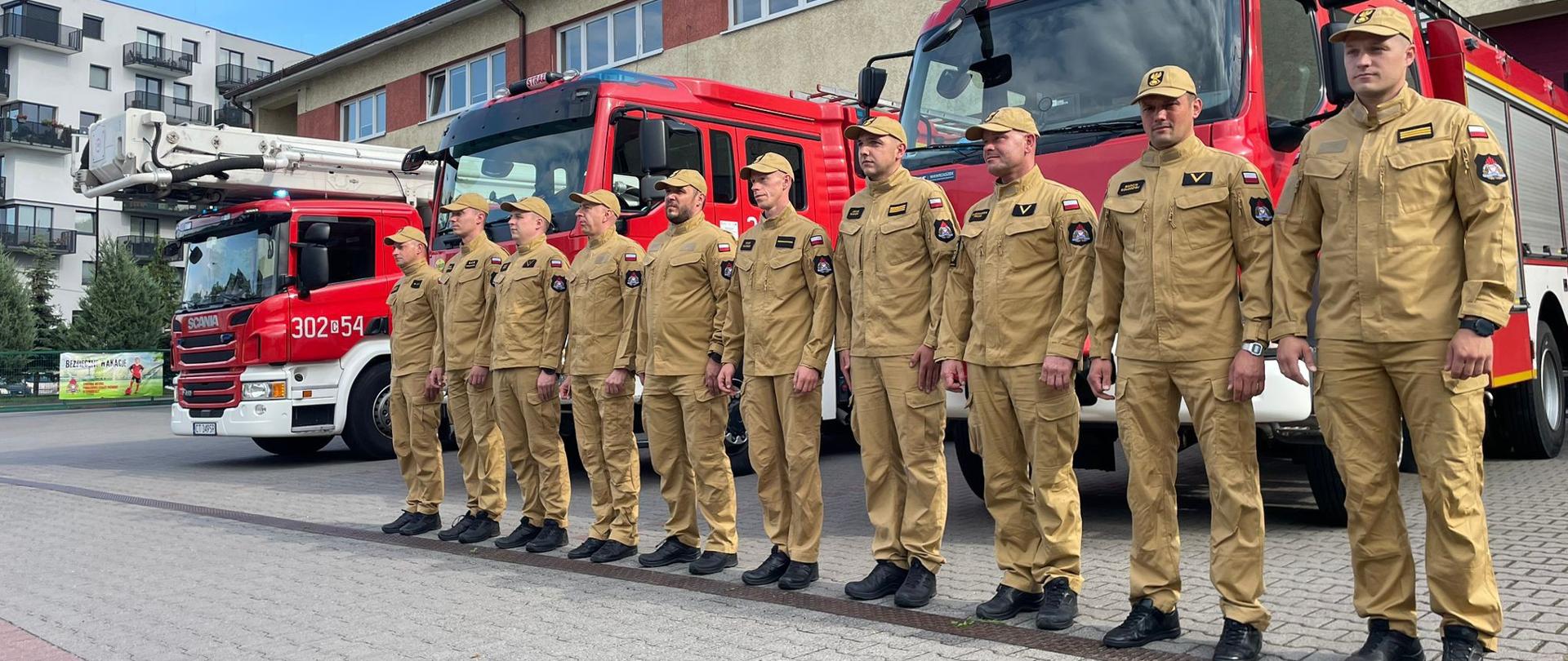 Zbiórka przed budynkiem JRG2 w Bydgoszczy. Strażacy w szeregu w żółtych ubraniach służbowych. Za nimi przody trzech pojazdów i elewacja frontowa budynku w żółtym kolorze i czerwonym paskiem wzdłuż okien. 