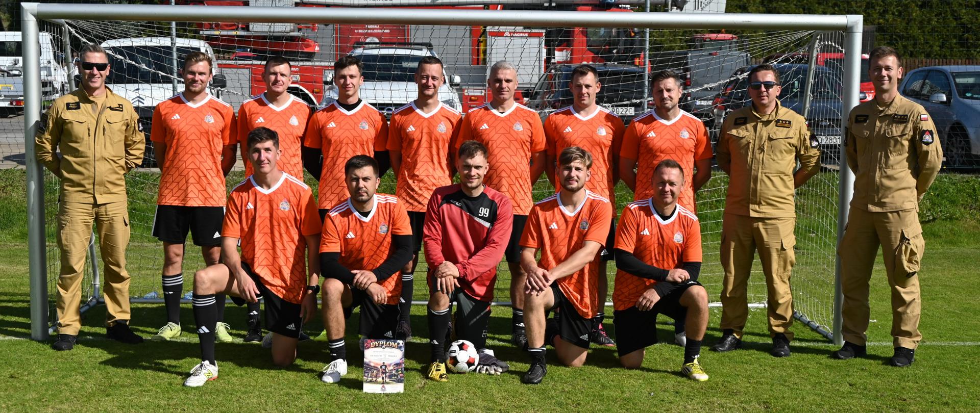 Grupa zawodników w pomarańczowo-czarnych strojach sportowych oraz bramkarz w czerwonej bluzie pozują do zdjęcia wewnątrz bramki piłkarskiej na boisku. Przed zawodnikami leży dyplom oraz piłka. Po obu stronach bramki stoją strażacy w piaskowych mundurach służbowych. W tle za siatką widoczny jest wóz strażacki z drabiną Magirus oraz zaparkowane samochody.