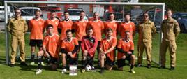 Grupa zawodników w pomarańczowo-czarnych strojach sportowych oraz bramkarz w czerwonej bluzie pozują do zdjęcia wewnątrz bramki piłkarskiej na boisku. Przed zawodnikami leży dyplom oraz piłka. Po obu stronach bramki stoją strażacy w piaskowych mundurach służbowych. W tle za siatką widoczny jest wóz strażacki z drabiną Magirus oraz zaparkowane samochody.