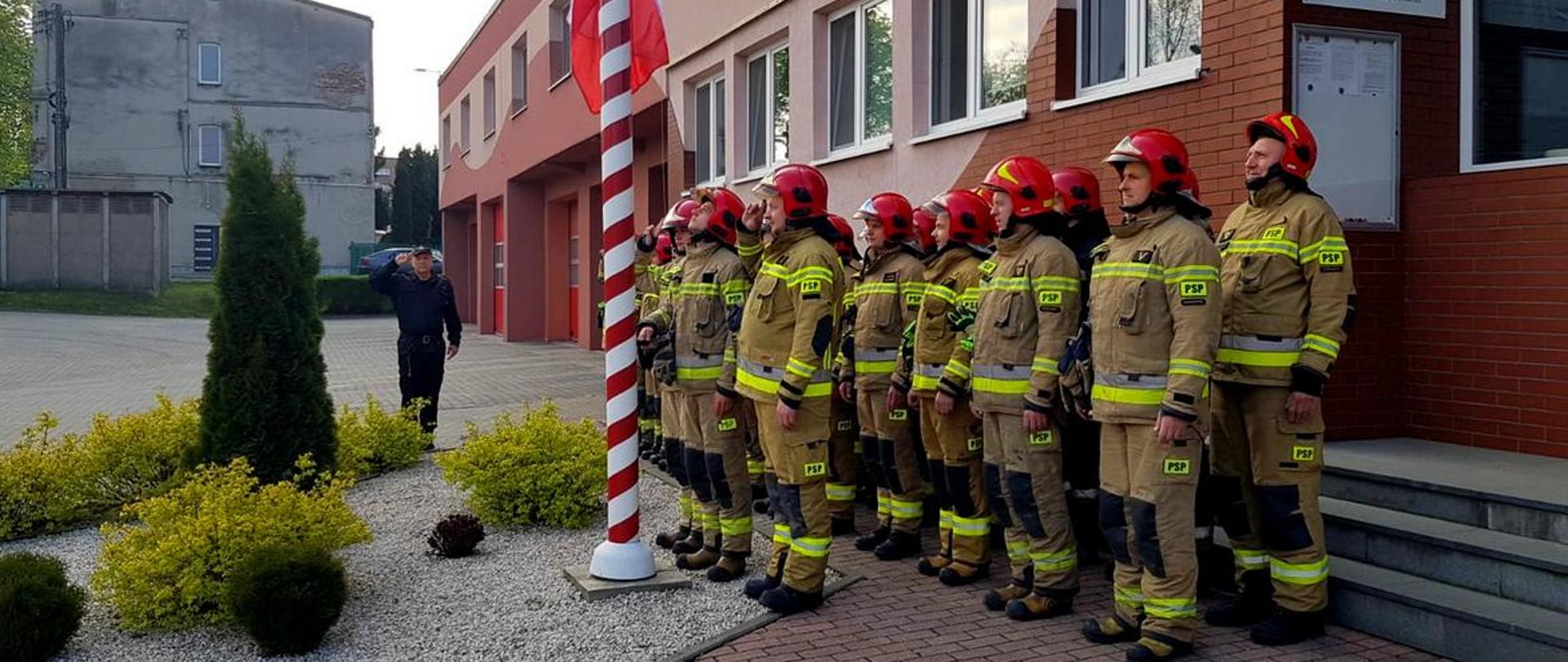 Na zdjęciu funkcjonariusze w ubraniu specjalnym oraz Dowódca Jednostki Ratowniczo-Gaśniczej ubrany w ubranie dowódczo sztabowe podczas uroczystej zmiany służby, podczas której miało miejsce uroczyste podniesienie flagi państwowej. 