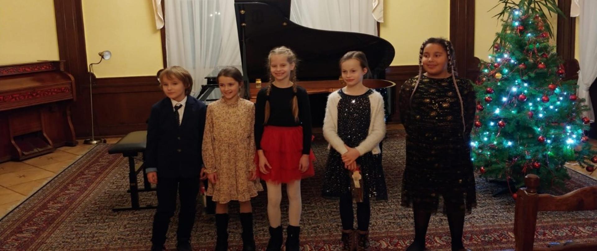 Zdjęcie kolorowe. Uczestnicy konkursu - dzieci w strojach galowych stoją na tle fortepianu w sali kameralnej Państwowej Szkoły Muzycznej I i II stopnia w Słupsku. Z prawej strony choinka.