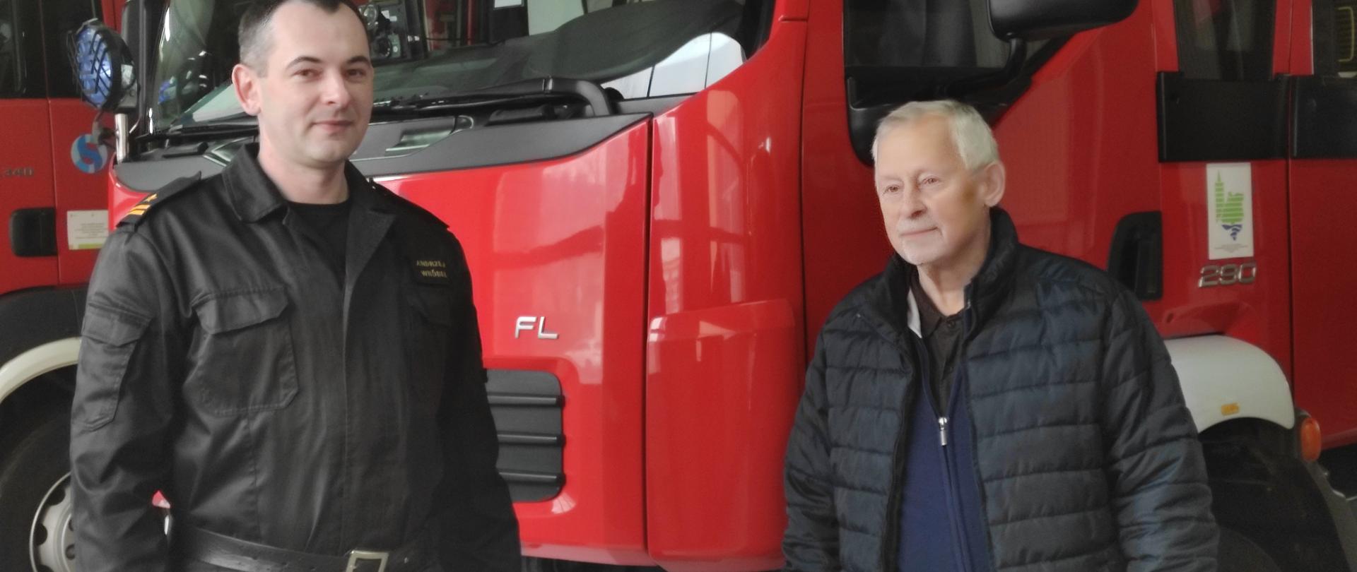 Strażak w ubraniu koszarowym stoi wraz ze starszym mężczyzną w ubraniu cywilnym w tle samochody pożarnicze.