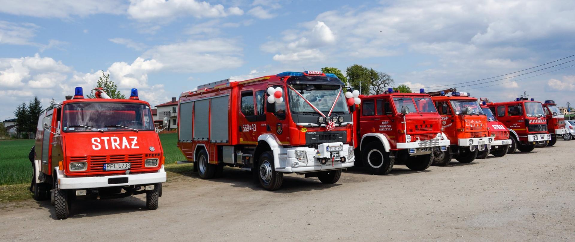 Wozy strażacki stojące w szeregu, drugi wóz od lewej jest udekorowany wstęgami i balonami