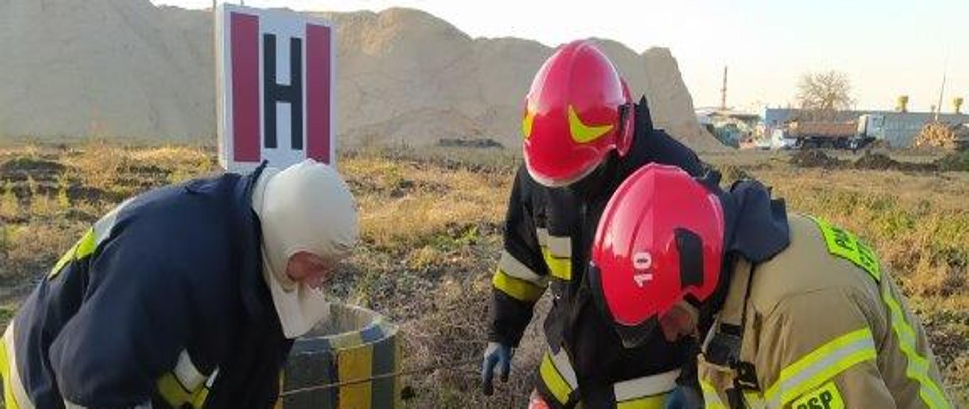 Na zdjęciu trzech strażaków pochylonych nad otwartą studzienką kanalizacyjną. Za plecami strażaka z lewej znak H oznaczający hydrant. W tle hałda piasku. 