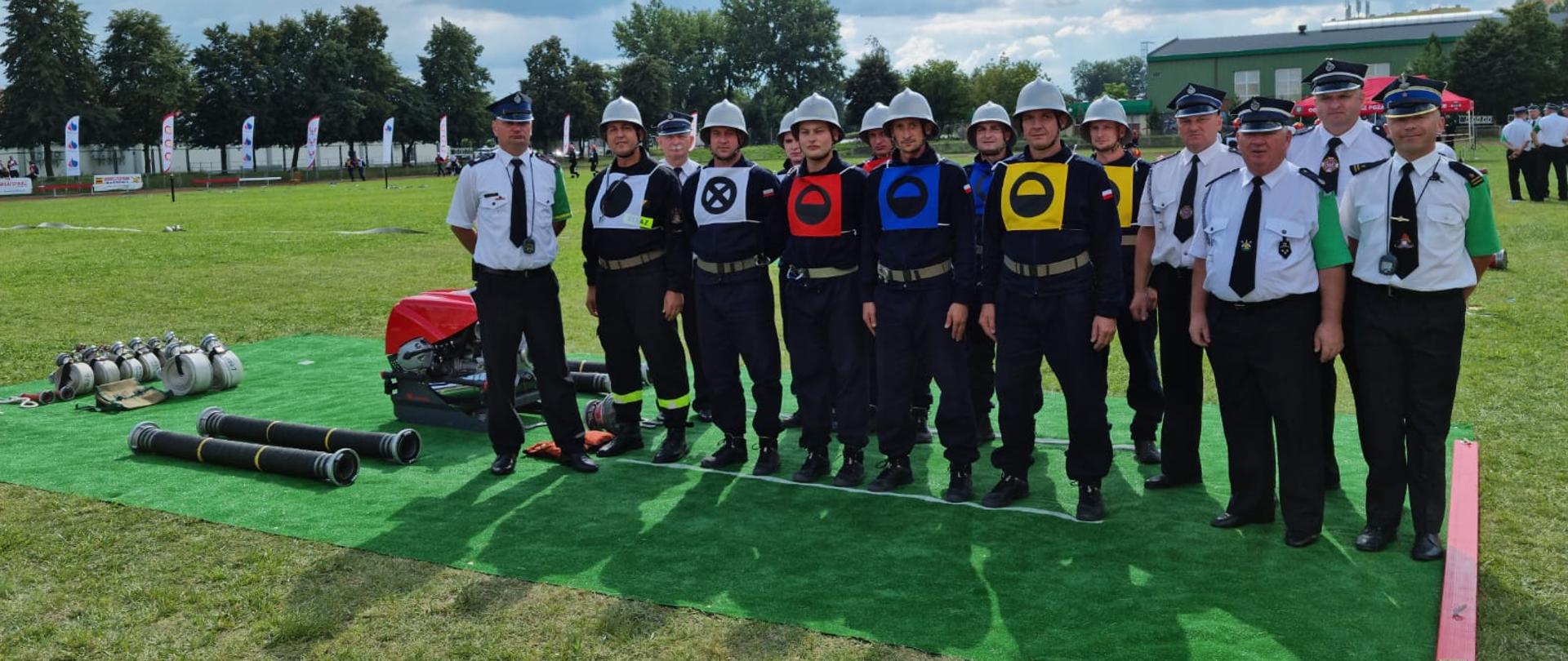 Drużyna sportowa KP PSP w Pleszewie ustawiona w dwuszeregu, przed startem. Wraz z zawodnikami stoją sędziowie zawodów, obok widoczny jest sprzęt do ćwiczeń.