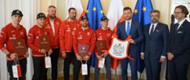 Minister Bortniczuk spotkał się z reprezentantami Polski, drużynowymi mistrzami Europy. Na zdjęciu minister pozuje z otrzymanym od żużlowców plastronem. Obecni na spotkaniu żużlowcy to Bartosz Zmarzlik, Jarosłąw Hampel, Szymon Woźniak i Bartłomiej Kowalski, którzy trzymają w rękach listy gratulacyjne i upominki od ministra. Na zdjęciu widoczni także przedstawiciele Polskiego Związku Motorowego. Cała grupa osób stoi na tle flag Polski i Unii Europejskiej. Żużlowcy ubrani są w czerwone bluzy z orzełkiem na piersi.