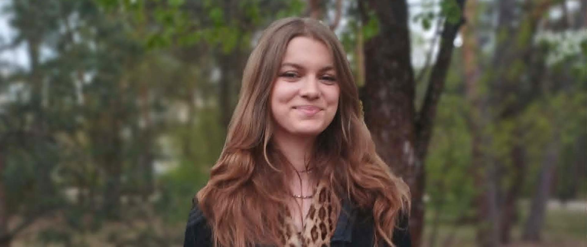 Młoda kobieta z długimi brązowymi włosami, ubrana w czarną marynarkę i sukienkę, stoi na zewnątrz w lesie lub parku, uśmiecha się do kamery i trzyma przed sobą dyplom z czerwono-białą grafiką czołgu i napisem „DYPLOM”. Tło jest lekko rozmyte, z drzewami.