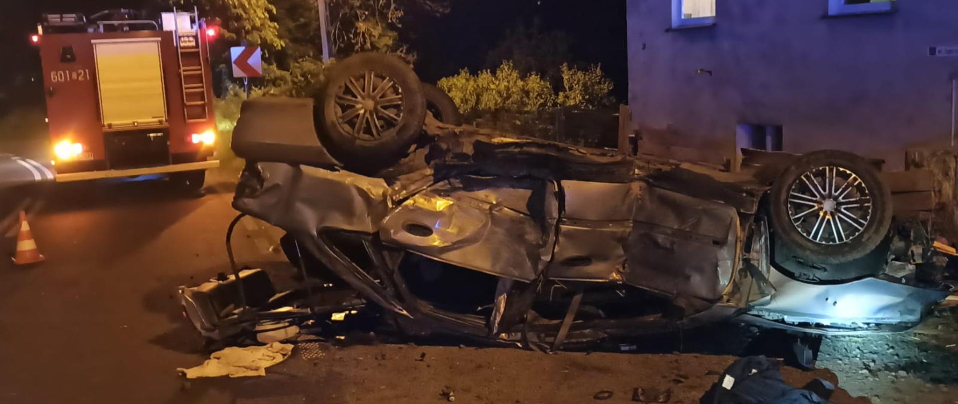 Pora nocna. Samochód osobowy na dachy na poboczu drogi. Z prawej strony budynek mieszkalny. Z lewej strony w tle pojazd ratowniczo -gaśniczy.