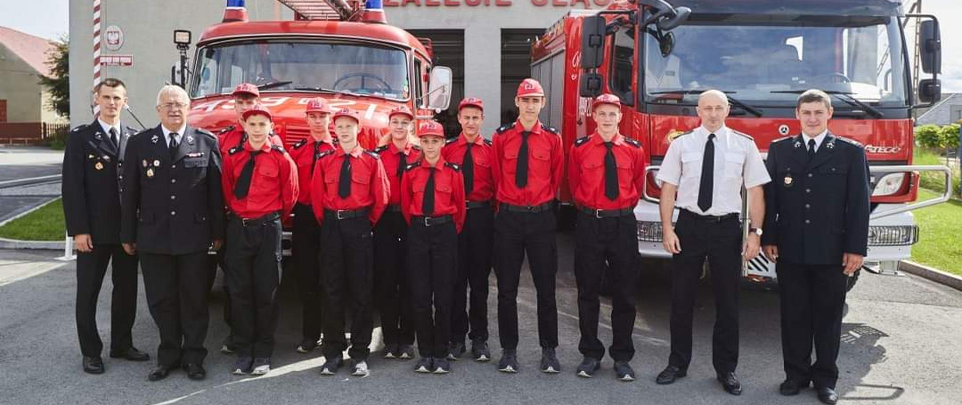 Na zdjęciu znajdują się strażacy oraz członkowie MDP i Zastępca OKW w mundurach ustawieni na tle samochodów pożarniczych oraz remizy.