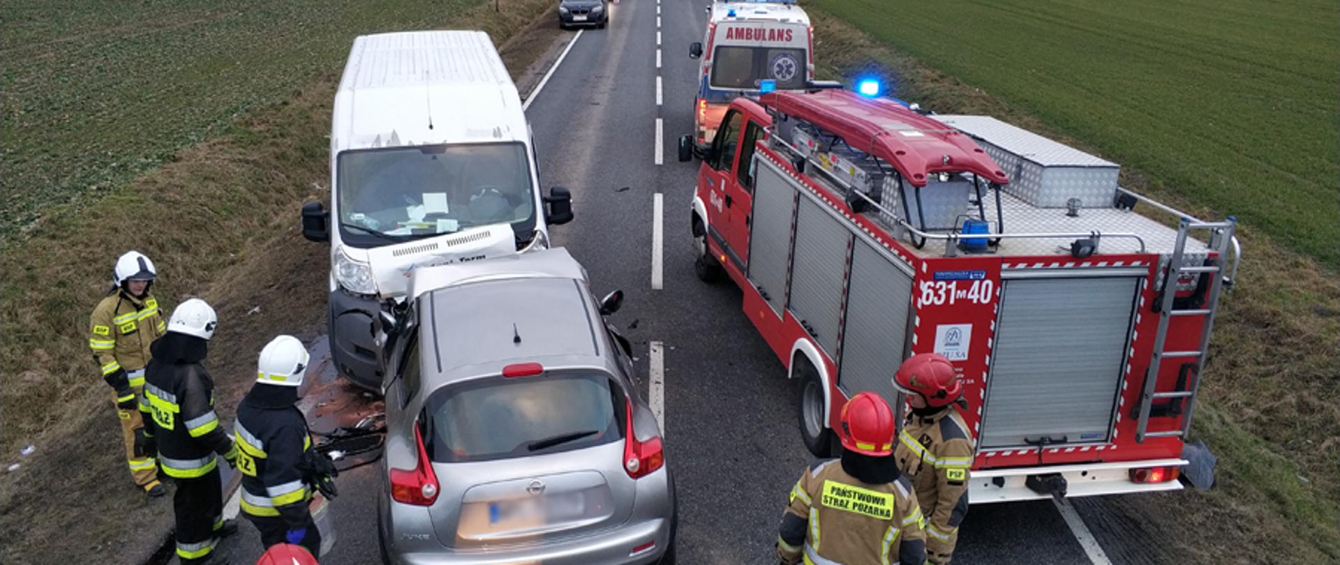 Na zdjęciu widoczny srebrny samochód osobowy, który zderzył się czołowo z białym busem. Widać samochód straży pożarnej oraz ambulans. Widać także trzech strażaków w białych hełmach i trzech w czerwonych.