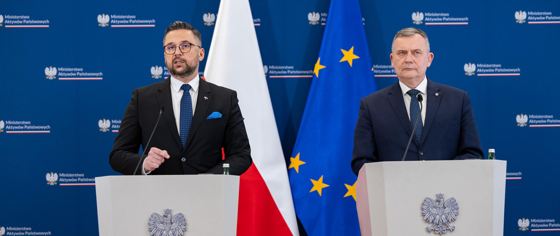 Dwaj mężczyźni (Marcin Kulasek, wiceminister aktywów państwowych oraz Paweł Bejda, wiceminister obrony narodowej) w garniturach stoją przy mównicach z orłem i napisem Ministerstwo Aktywów Państwowych. W tle ścianka z logotypem resortu. Po środku flagi (od lewej): Polski i UE.