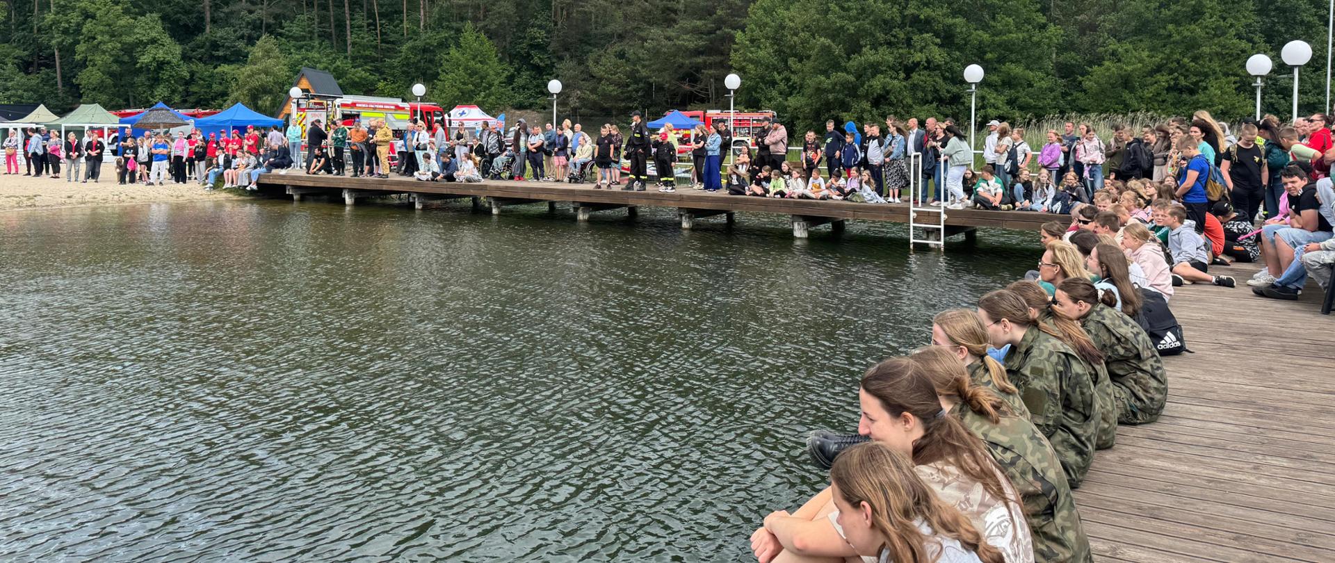 Na zdjęciu widzimy uczestników akcji "Bezpieczne wakacje" siedzących na pomoście nad jeziorem