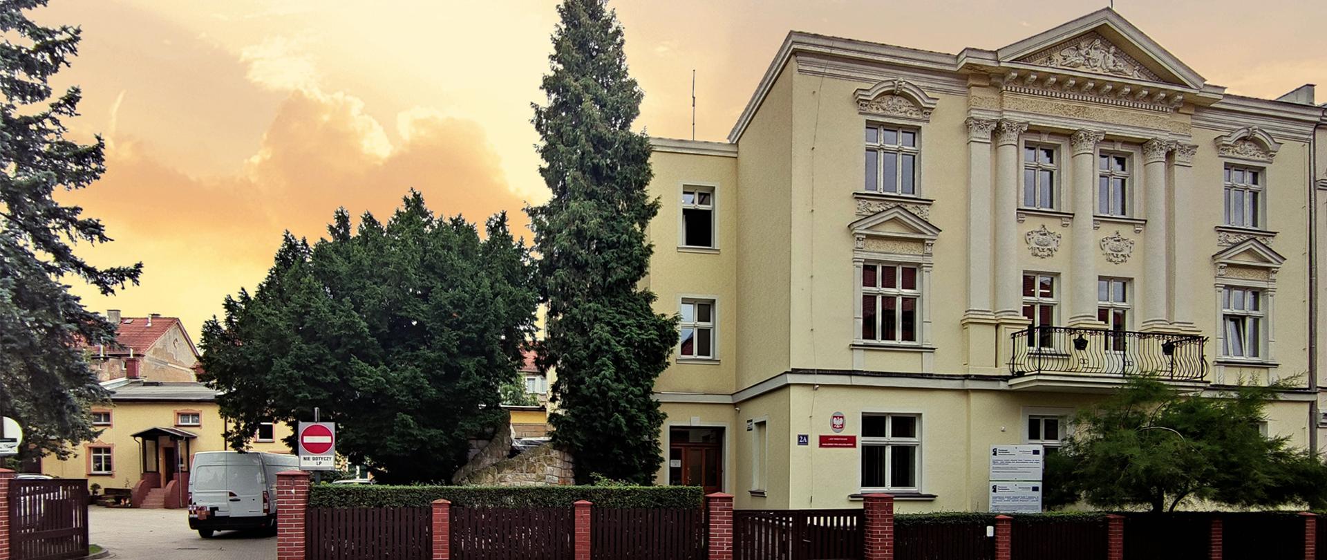 widok budynku siedziby Nadleśnictwa Bolesławiec o zachodzie słońca od strony ulicy. Budynek umiejscowiony jest po prawej stronie obrazka a jego centralną część zajmują drzewa pod którymi znajduje się wjazd na plac przy nadleśnictwie oraz zaparkowane na nim samochody. 