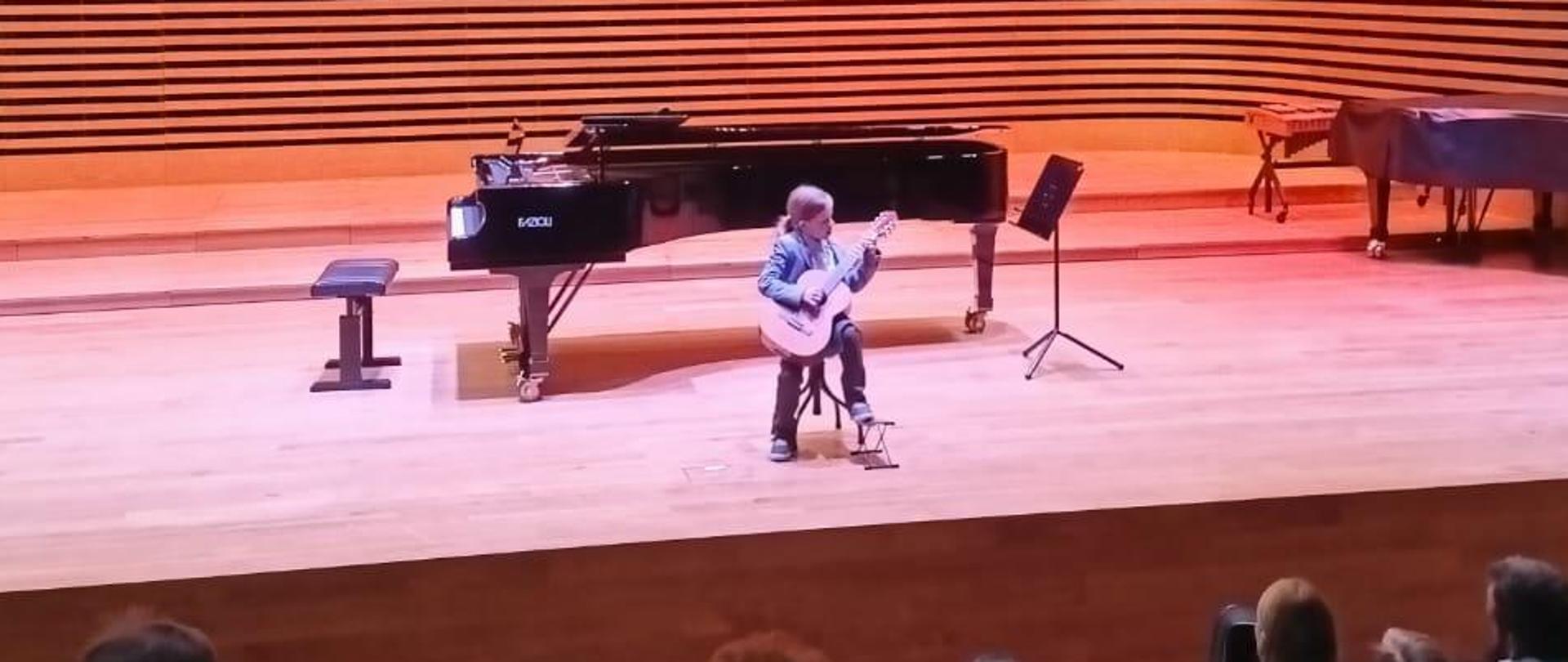 Chłopiec gra na gitarze, za Nim stoi fortepian.