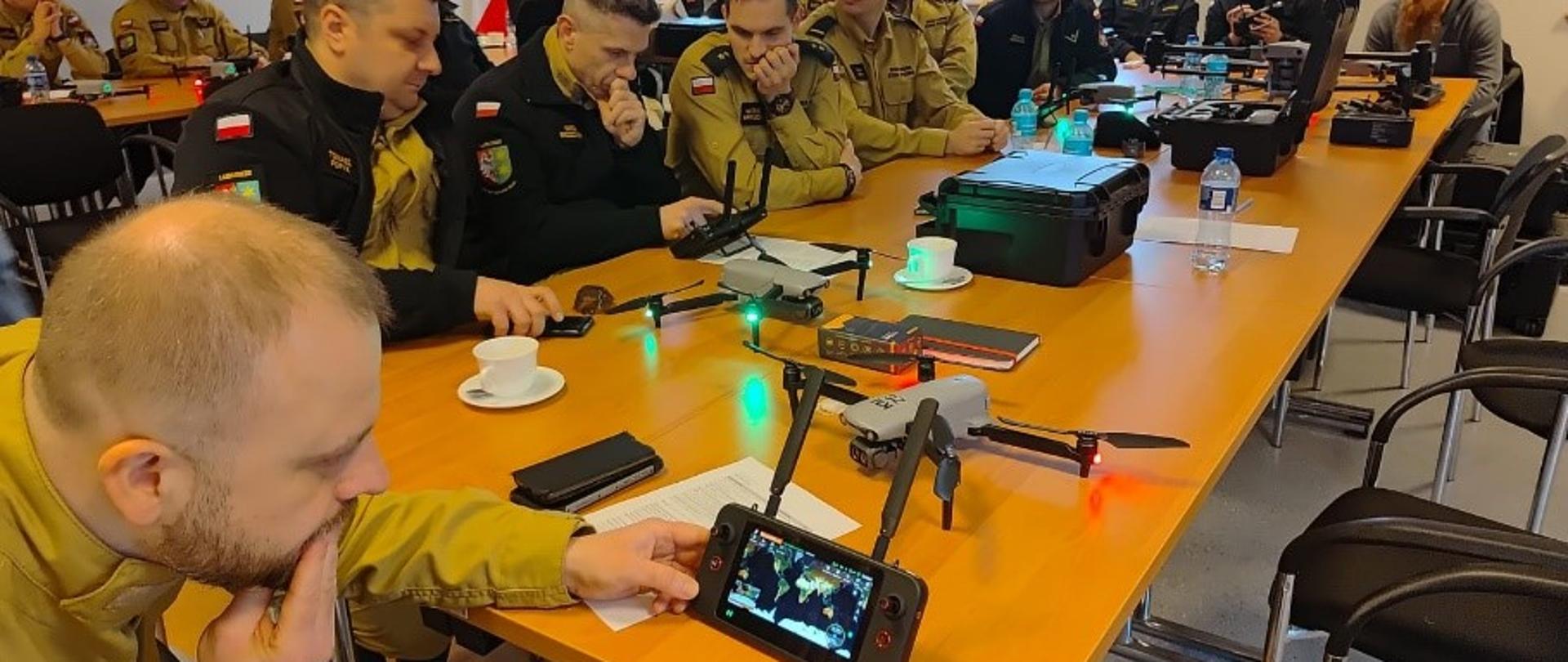 Sala szkoleniowa, przy długim stole siedzi grupa strażaków w mundurach PSP, uważnie przyglądających się kontrolerowi drona trzymanemu przez jednego z nich. Na stole znajdują się rozłożone drony, walizki transportowe oraz filiżanki z kawą i notatniki.