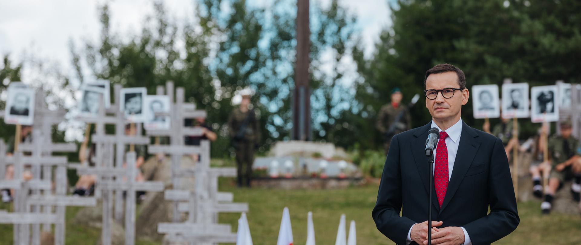 Premier stoi przed pomnikiem. W tle polska flaga oraz drewniane krzyże symbolizujące pomordowanych przez NKWD partyzantów niepodległościowych.