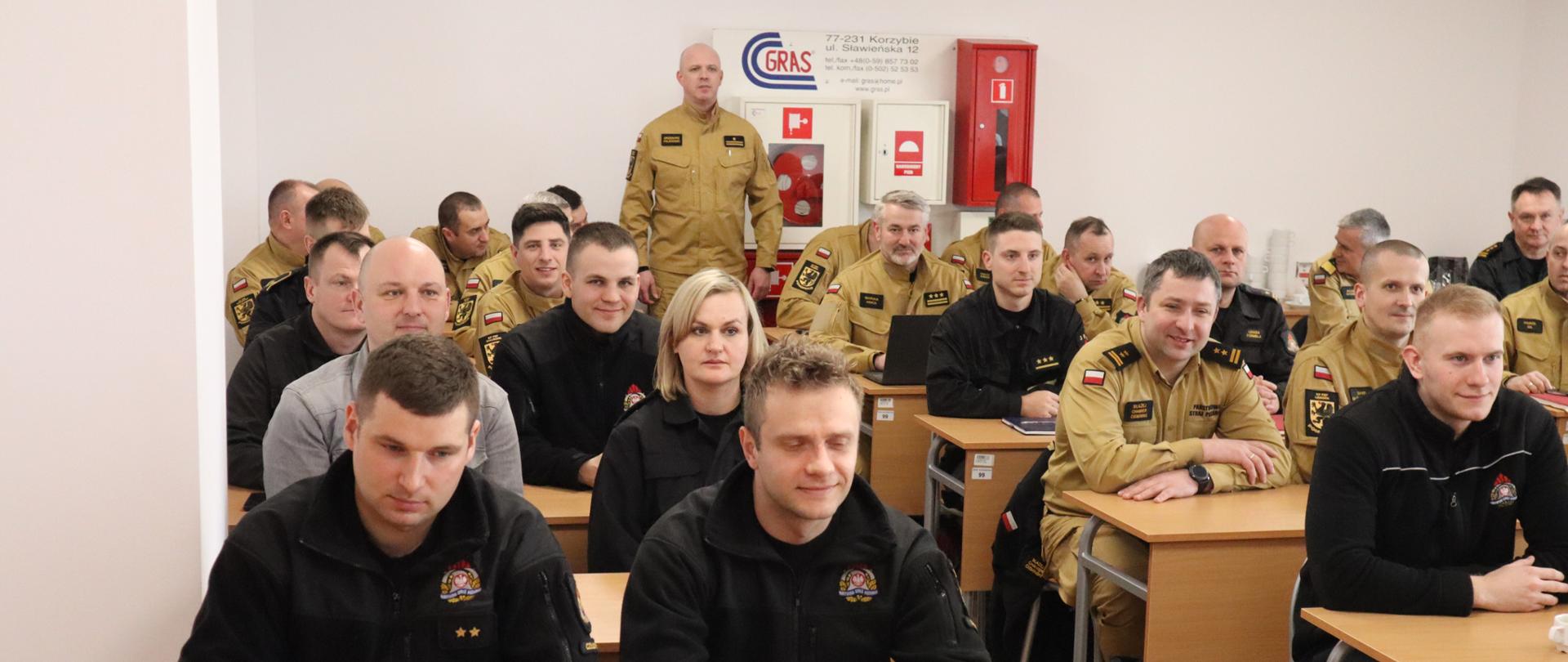 Funkcjonariusze Państwowej Straży Pożarnej oraz pracownicy cywilni siedzą w ławkach podczas szkolenia. Strażacy ubrani są w mundury w kolorze czarnym lub piaskowym. 
