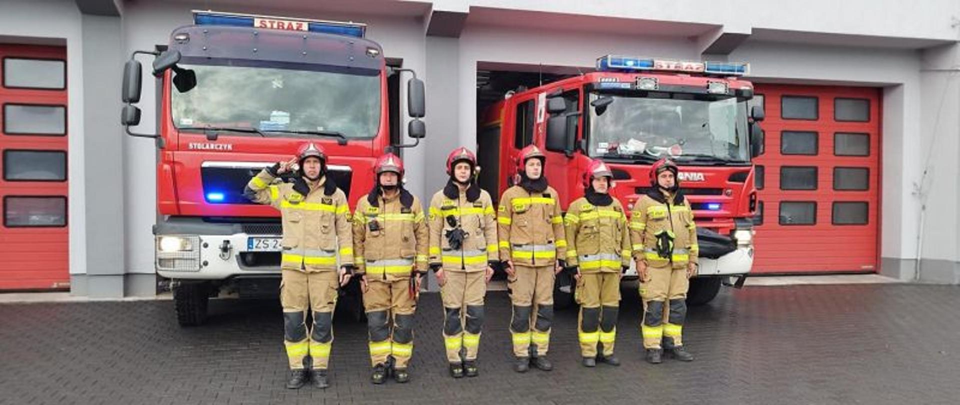 Zdjęcie wykonane przed budynkiem komendy. W tle bramy i pojazdy gaśnicze. Na pierwszym planie w szeregu strażacy w żółtych mundurach.