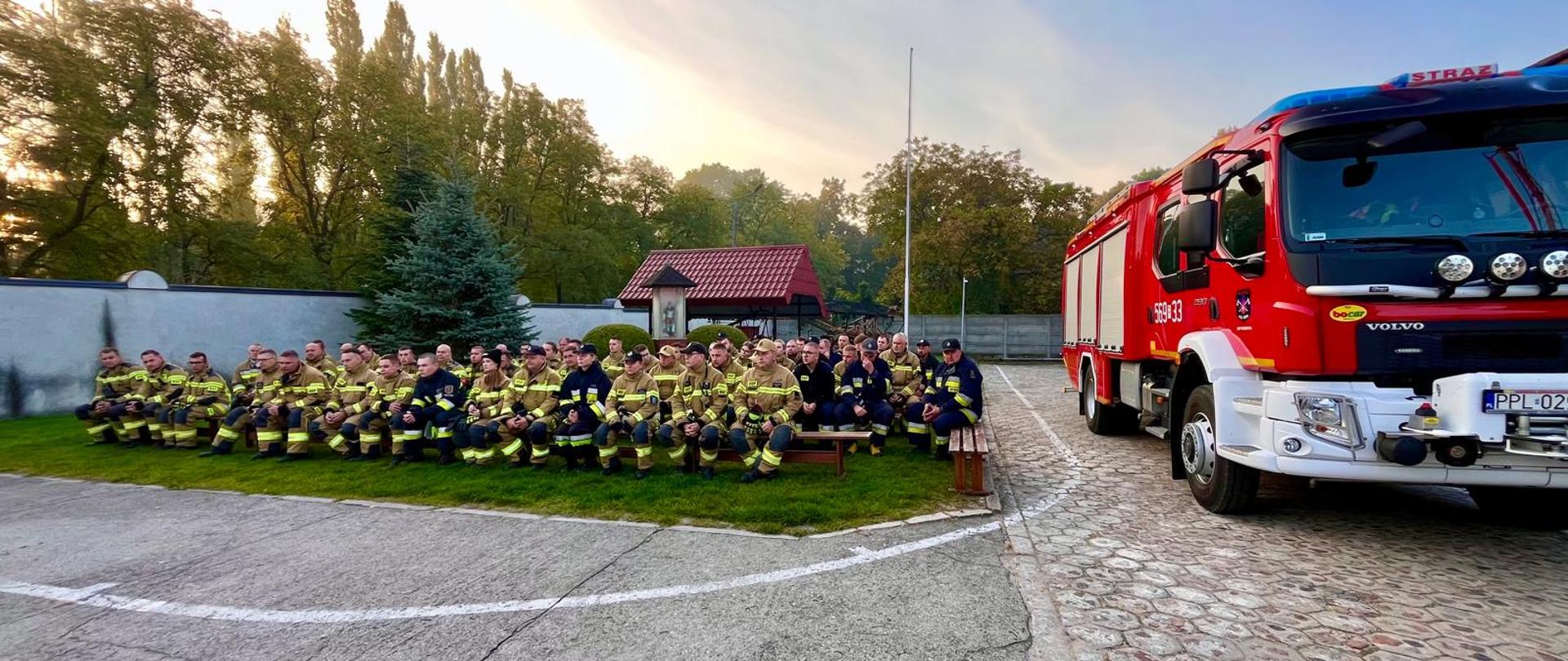 Zbiórka strażaków w mundurach bojowych, którzy siedzą na ławkach i trawie na zewnątrz, obok zaparkowanego nowoczesnego, czerwonego wozu ratowniczo-gaśniczego