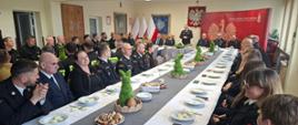 Przy stołach na których znajdują się dekoracje świąteczne i talerze, siedzą strażacy i zaproszenie goście. Strażacy ubrani są w mundury galowe zaproszeni goście odświętnie w garniturach. W tle logo Państwowej Straży Pożarnej oraz godło Polski i flagi