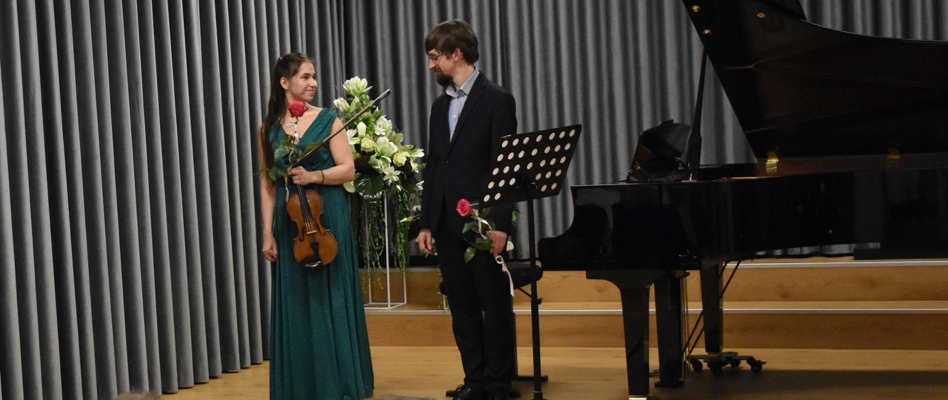 na zdjęciu, a uli PSM widnieją artyści (pianista i skrzypaczka). Na pierwszym planie widoczna jest społeczność szkolna, która oklaskuje artystów. Kolorystyka zdjęcia jest biało-szaro-brązowo-zielono-czarna.